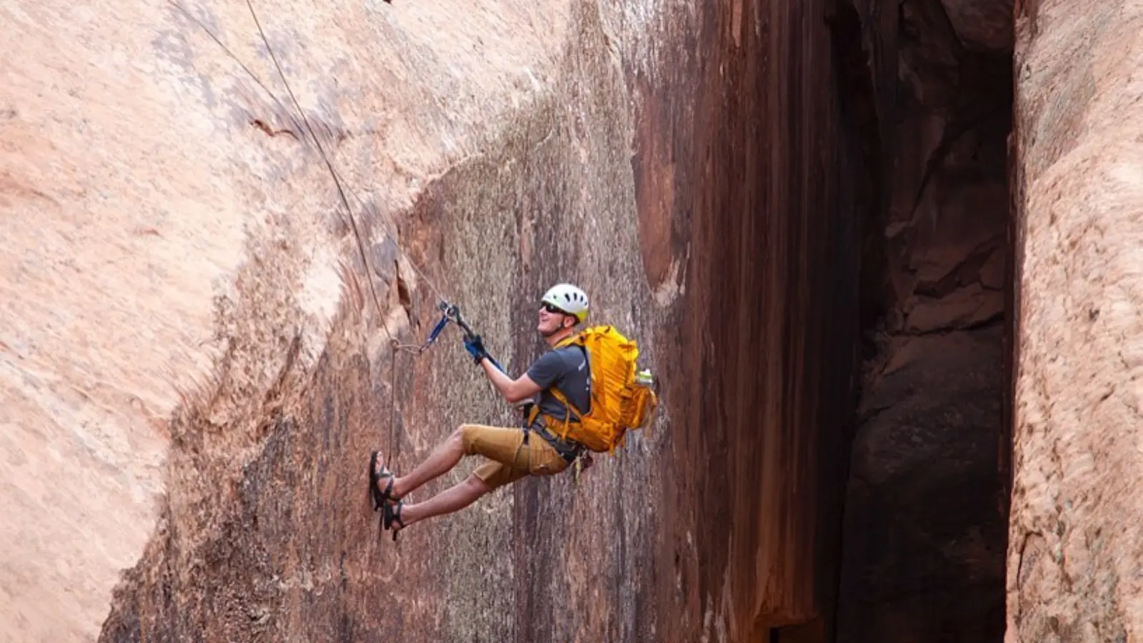 Five people in a canyon stand with helmets and climbing gear A man canyoneers down a rock wall in a yellow backapck
